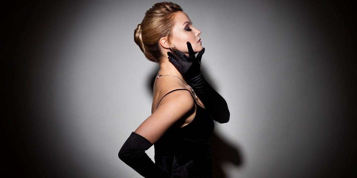 A femme fatale in a black dress with an open back and long velvet gloves. Attractive young blonde with makeup for a celebration, portrait in a spot of light